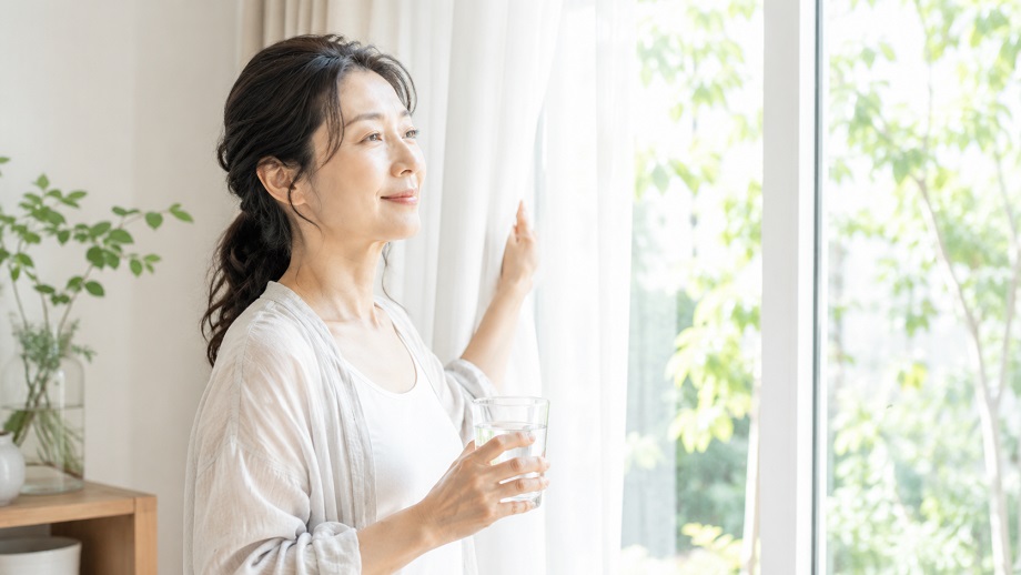朝の光の中でカーテンを開け、水を手に体を整える40代女性