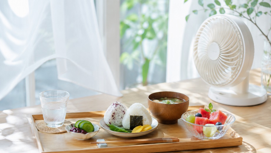 夏の明るい室内で、扇風機のそばに水、おにぎり、汁物、野菜、果物を並べた食卓