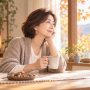 秋のやわらかな光が差し込む部屋で、マグカップを手に窓の外の紅葉を眺めながら穏やかにくつろぐ女性の姿。